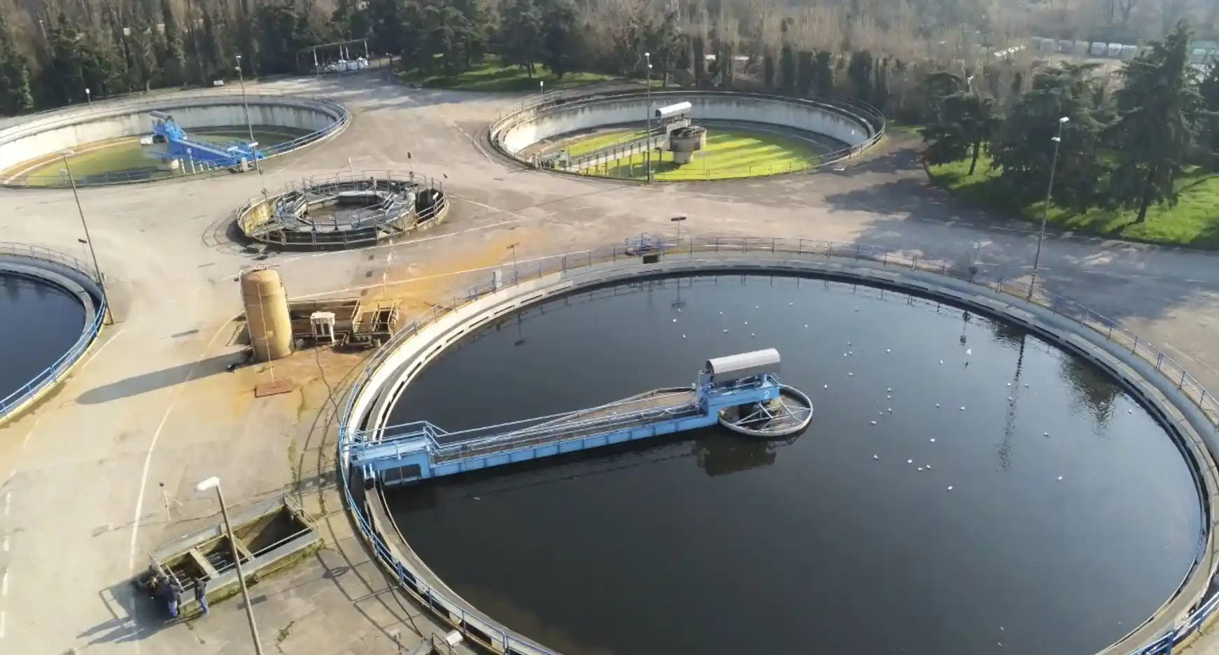 Questa è un'immagine aerea di un impianto di trattamento delle acque reflue. Ci sono diversi serbatoi rotondi, alcuni con acqua limpida e altri con acqua torbida, e una piattaforma con una passerella sopra uno dei serbatoi. L'impianto è circondato da alberi e vegetazione. L'impianto di trattamento delle acque reflue svolge un ruolo importante nel trattamento delle acque reflue prima che vengano restituite all'ambiente.