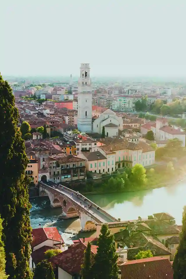 Uno sguardo aereo sulla città di Verona.  Il fiume Adige scorre attraverso la città, e il Ponte Scaligero attraversa il fiume. La torre campanaria di un'antica chiesa sorge sopra le case e gli alberi.  La città è un'incantevole miscela di architettura antica e moderna.