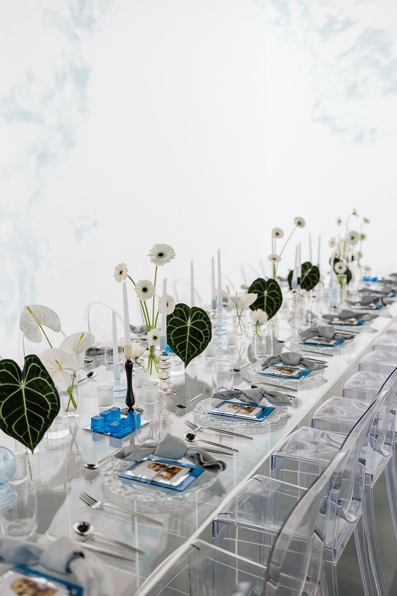A long, modern dining table is meticulously set for an event, featuring clear acrylic chairs and a reflective surface. T