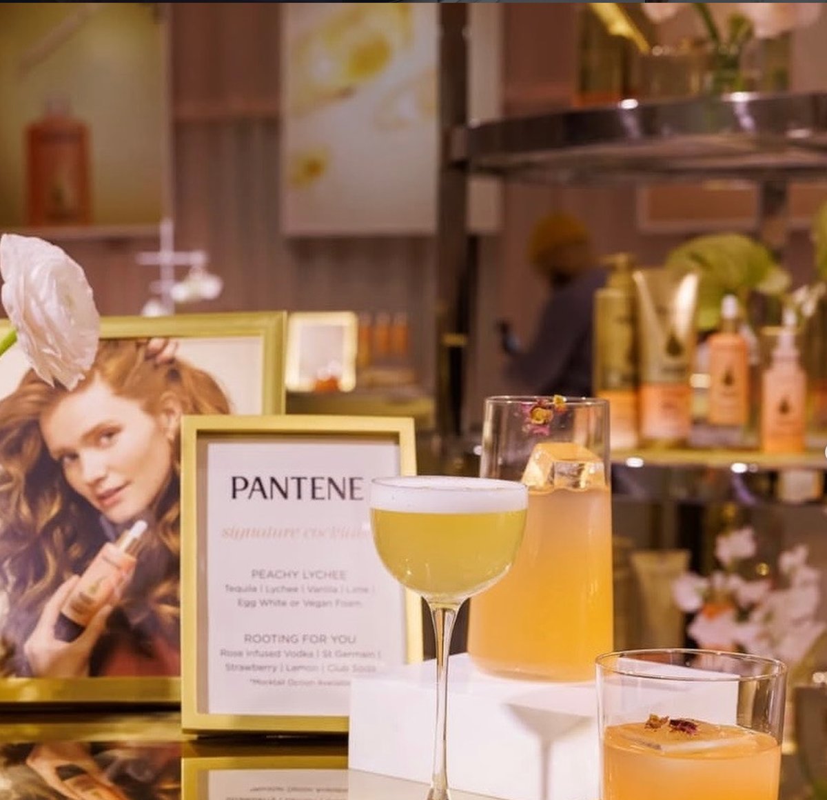 A close-up shot captures three vibrant cocktails displayed on a white stand, with a gold-framed Pantene advertisement an