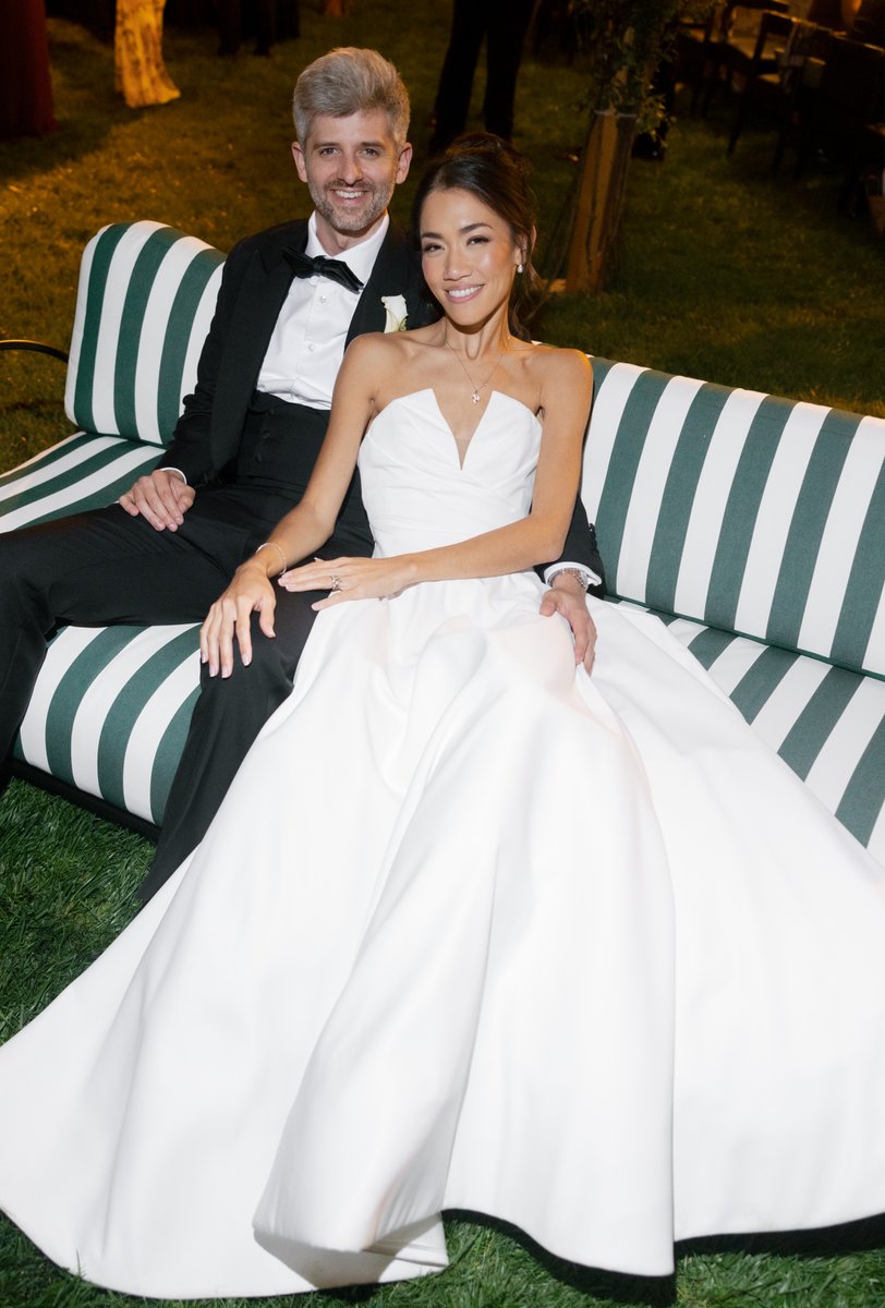 A happy couple, likely a bride and groom, is seated on a green and white striped outdoor bench. The woman wears a white 