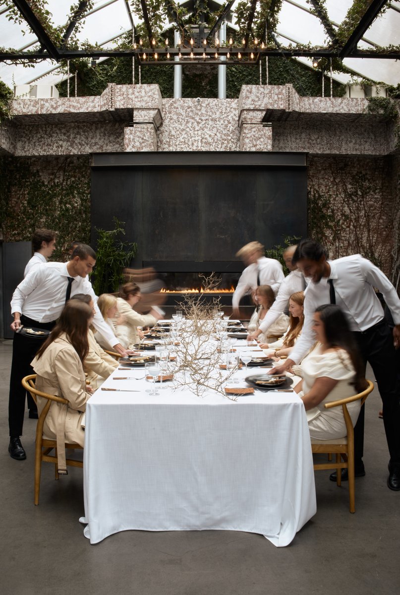 Servers in white shirts and black ties are actively attending to guests seated at a long, white-linened table in a moder