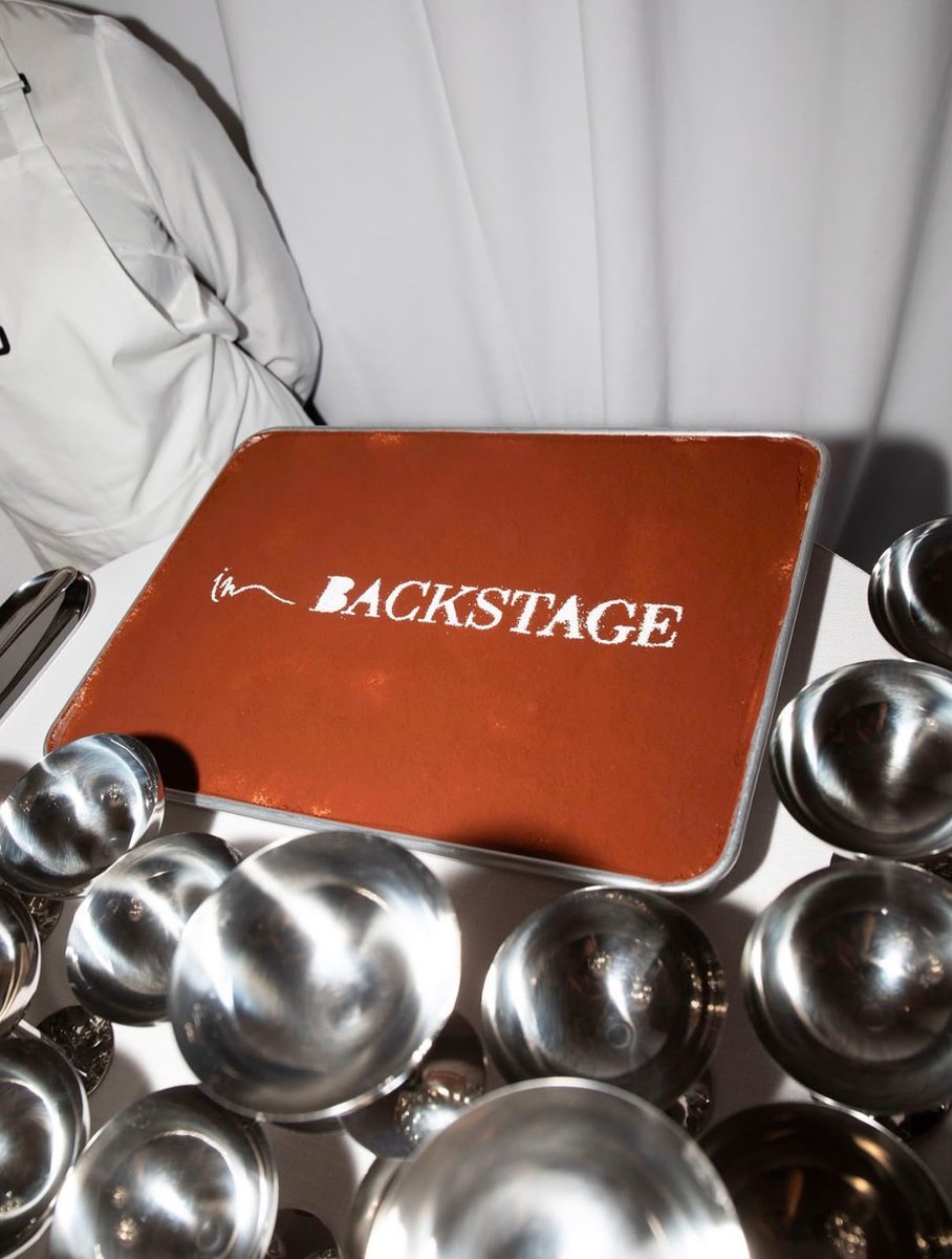 A chef, partially visible in a white uniform, presents a large rectangular dessert, possibly tiramisu, with "in BACKSTAG