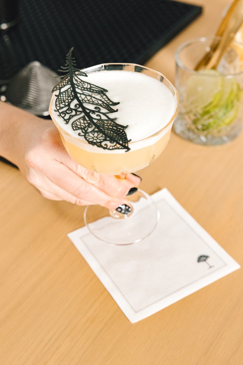 A stylish bartender with a man bun and beard is actively shaking a cocktail shaker behind a white bar, surrounded by ela