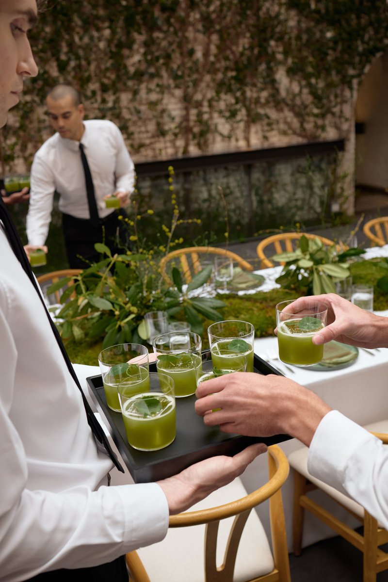 Two servers are actively serving refreshing green drinks with a leaf garnish at an outdoor event. One server in the fore