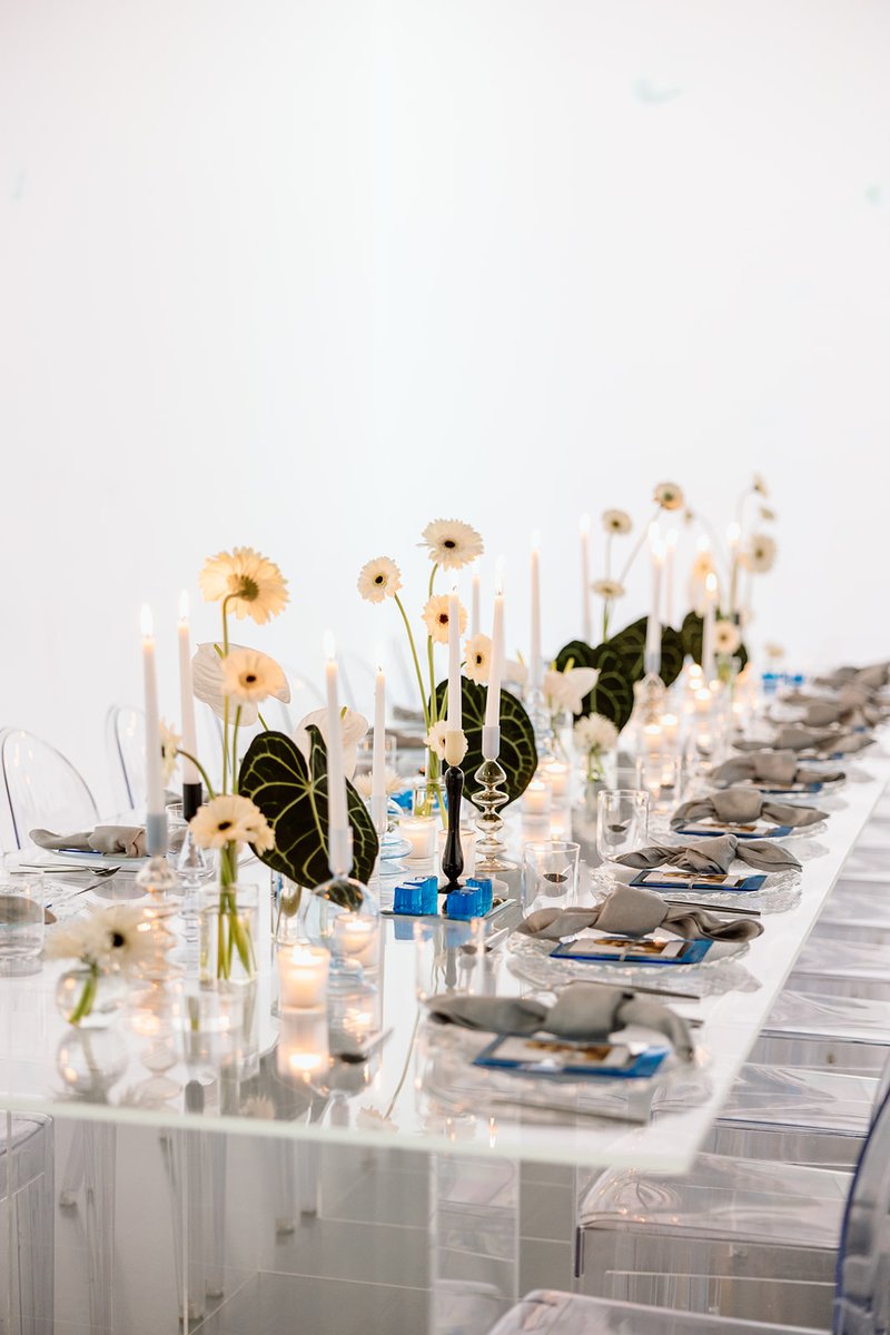 An elegantly set long dining table features white gerbera daisies, dark green anthurium leaves, and numerous white taper