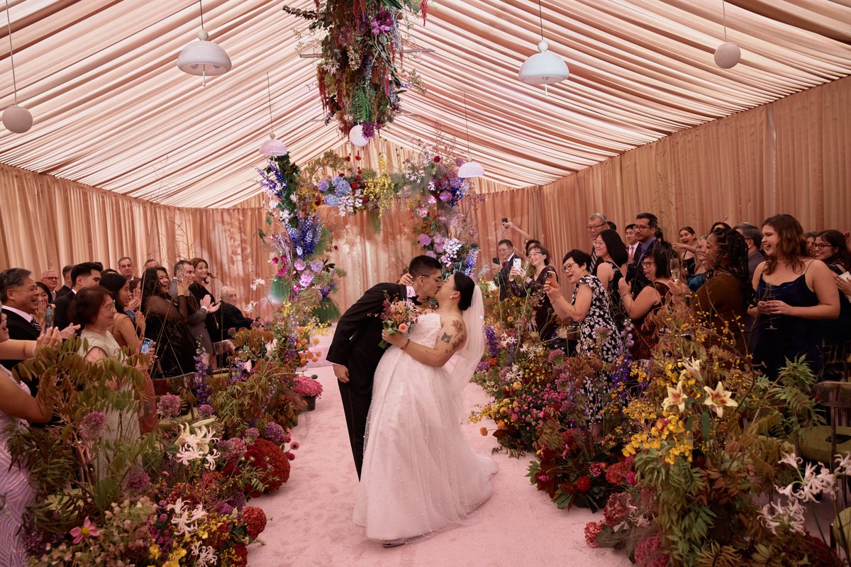 A newlywed couple shares a kiss on a pink carpet aisle, surrounded by an abundance of vibrant floral arrangements and an