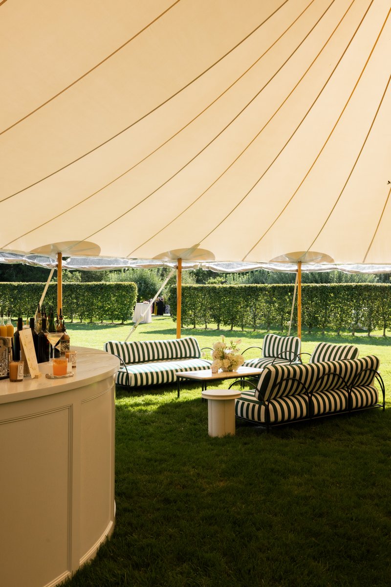 An elegant outdoor bar and lounge area is set up under a large cream-colored tent on a lush green lawn. A white curved b