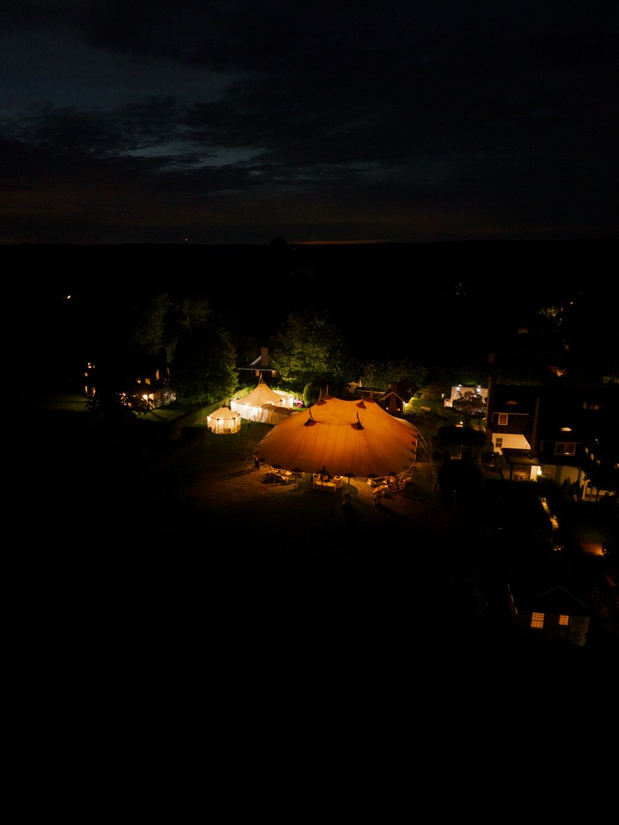 An aerial view captures a large, illuminated event tent at night, serving as the centerpiece of an outdoor gathering. Sm
