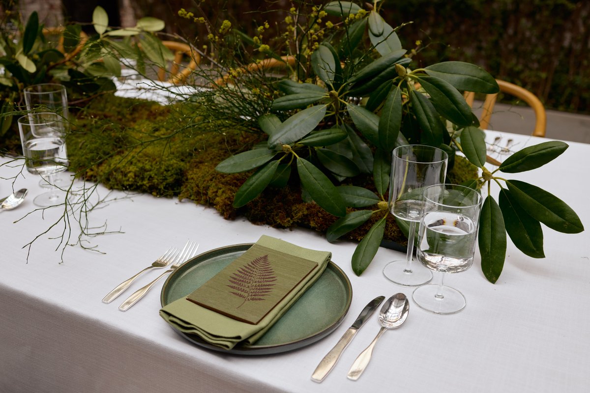 A beautifully set table features a lush, nature-inspired centerpiece made of moss and large green leaves. A single place