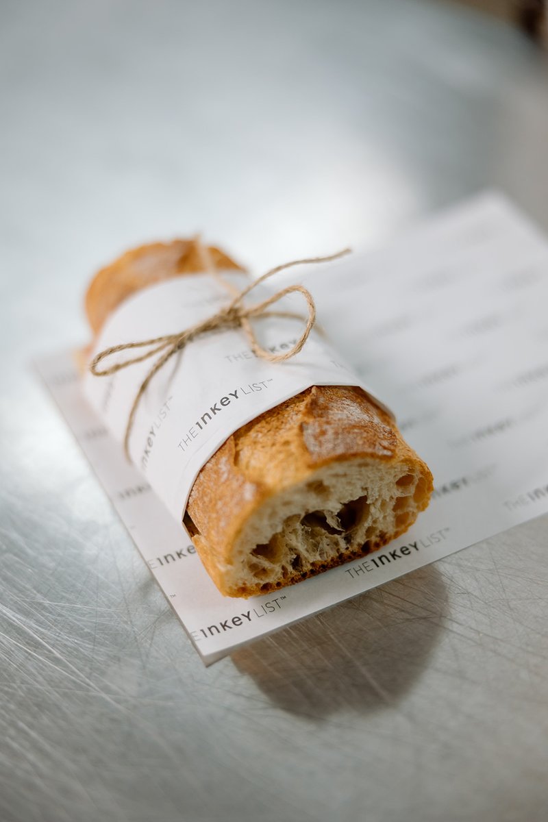 A freshly baked baguette, wrapped in white paper branded with 'THE INKEY LIST' and tied with rustic twine, rests on a li