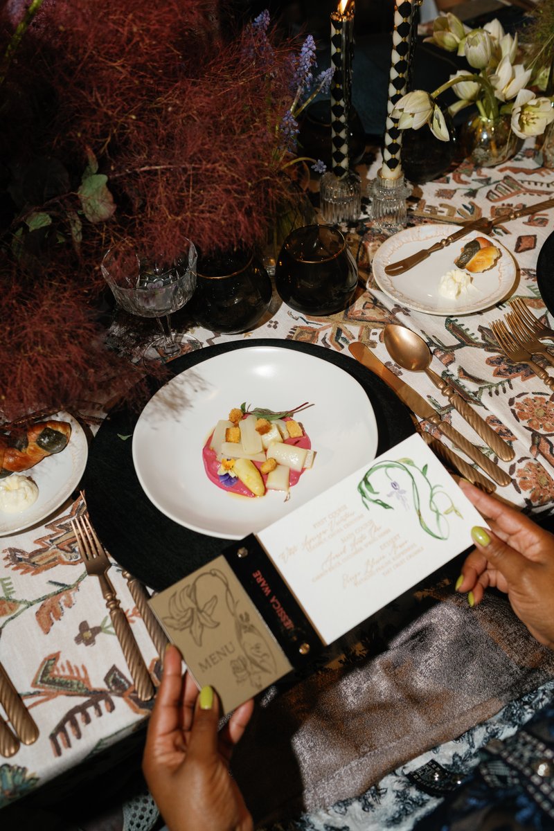 A close-up shot of an elegantly set dinner table, featuring a beautifully plated dish, ornate cutlery, and a guest's han