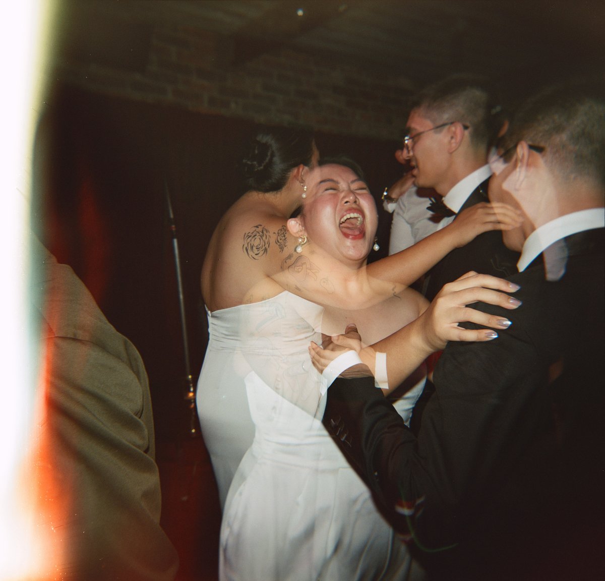 A group of guests, including a woman in a white dress, are captured mid-dance, laughing exuberantly at an indoor event. 