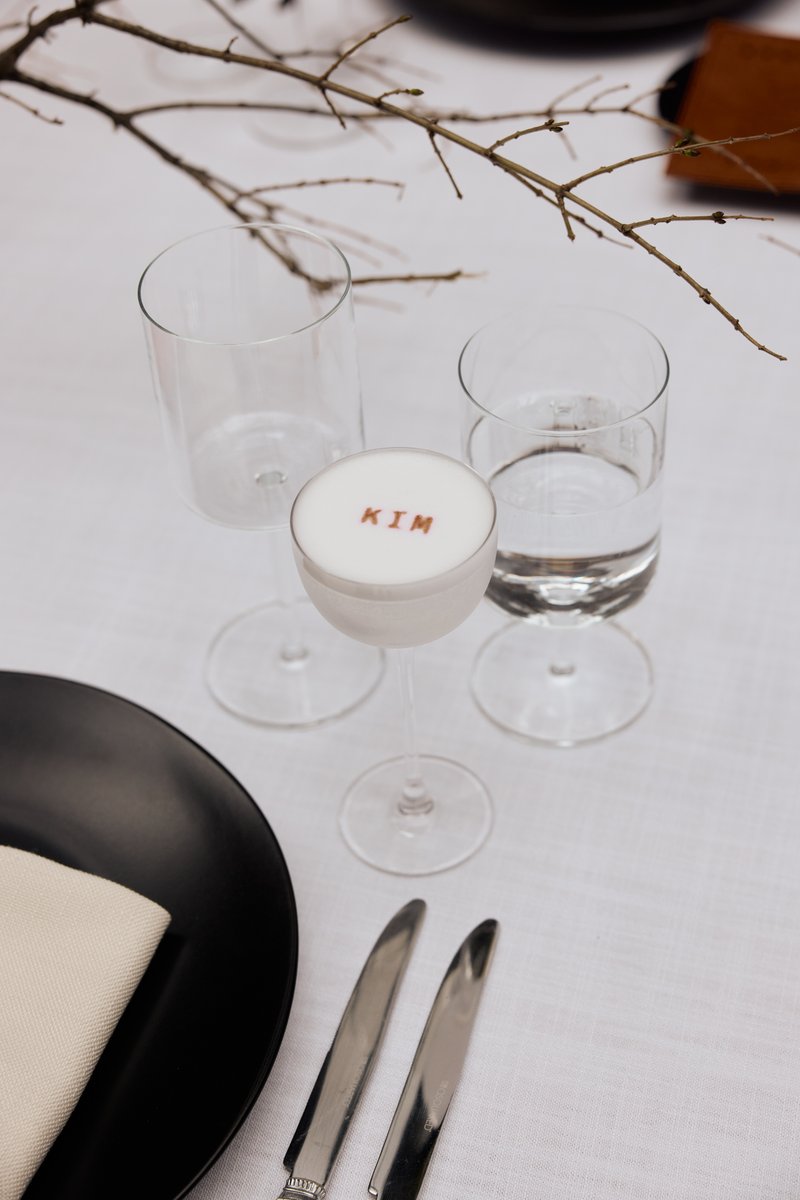 A minimalist and elegant table setting features a personalized cocktail with 'KIM' stenciled on its foam, alongside two 