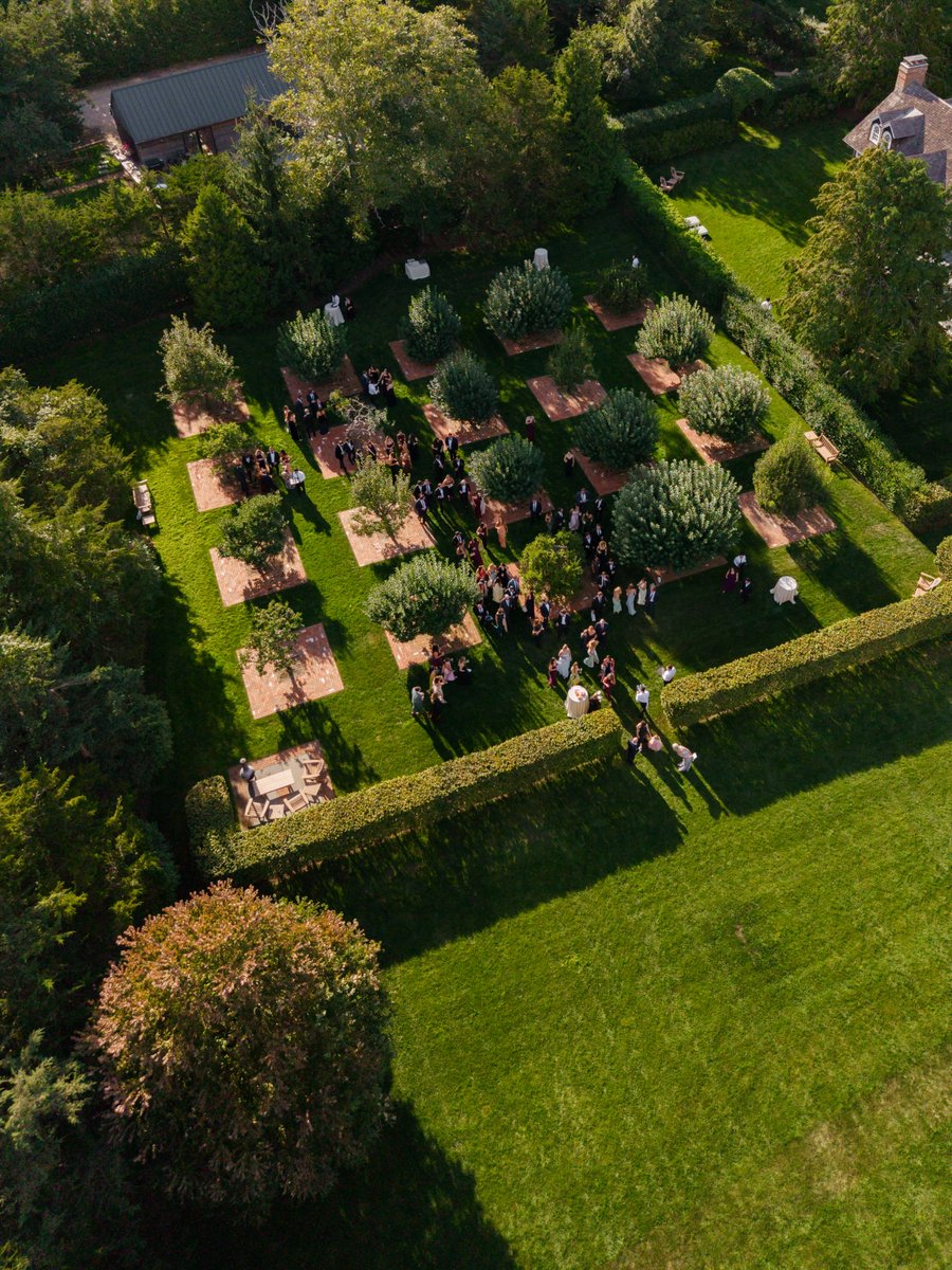 An aerial view captures a large outdoor event taking place in a beautifully manicured garden. Guests are seen mingling i