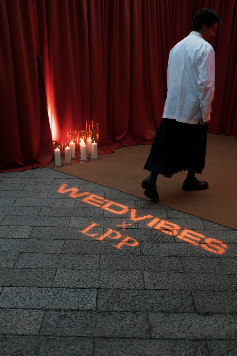 A person in a white shirt and dark skirt walks across a stone floor with an orange 'WEDVIBES x LPP' light projection. In