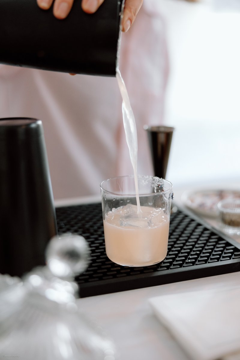 A close-up shot captures a bartender's hands expertly pouring a light pink cocktail from a black shaker into a salt-rimm