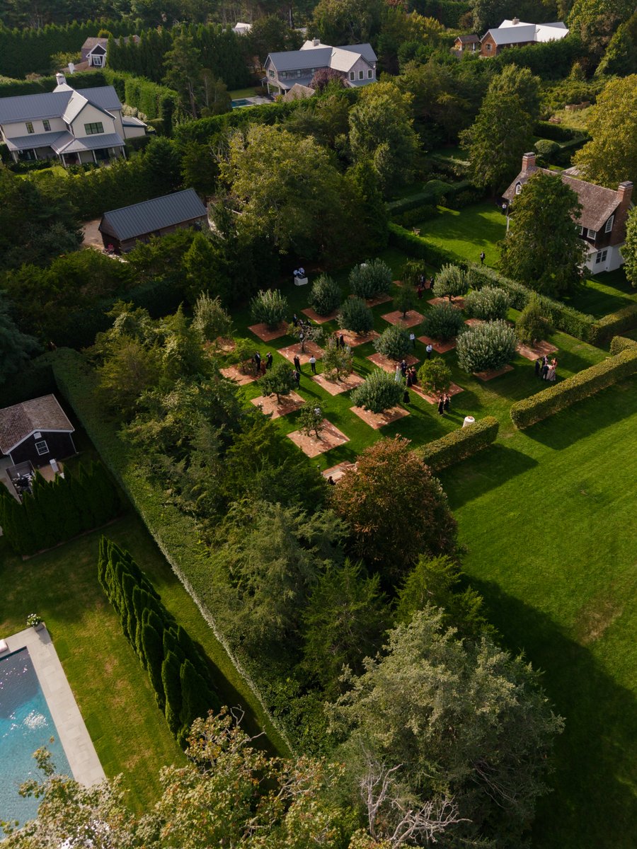 An aerial view captures an elegant outdoor event in a meticulously manicured garden. Guests are seen mingling among stru