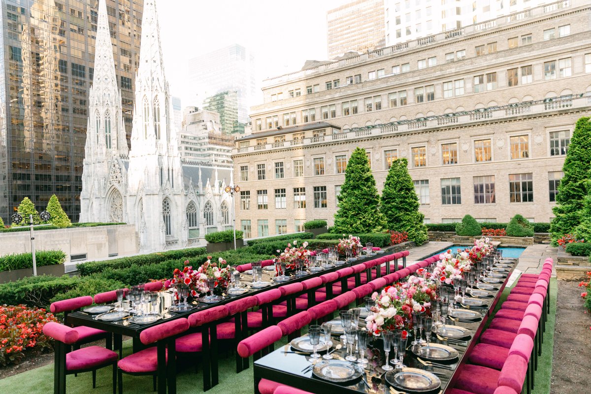 A beautifully set outdoor event space features two long tables adorned with vibrant pink chairs and lush floral arrangem