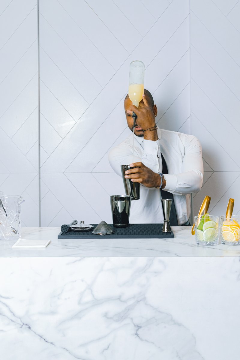 A professional bartender, dressed in a white shirt and dark tie, is expertly pouring a light yellow liquid from a bottle