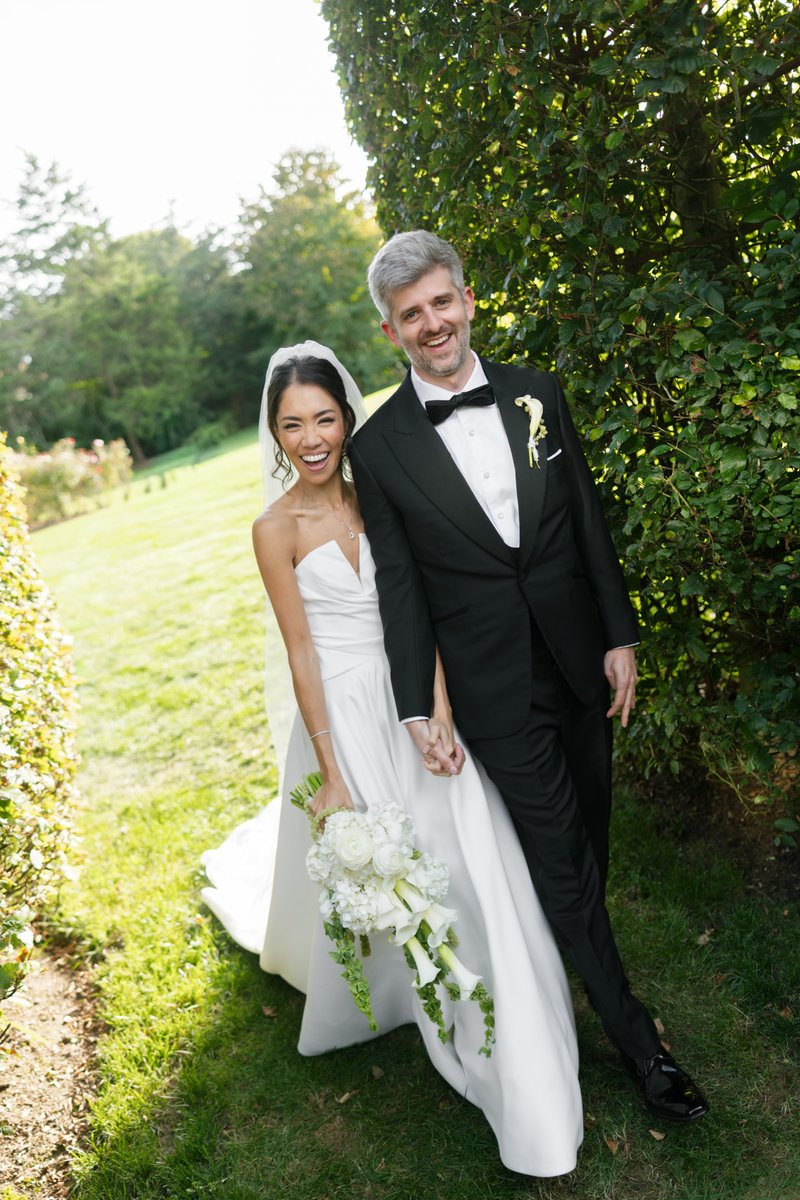 A happy bride and groom pose for a portrait outdoors, holding hands and smiling broadly. They are standing on a grassy p
