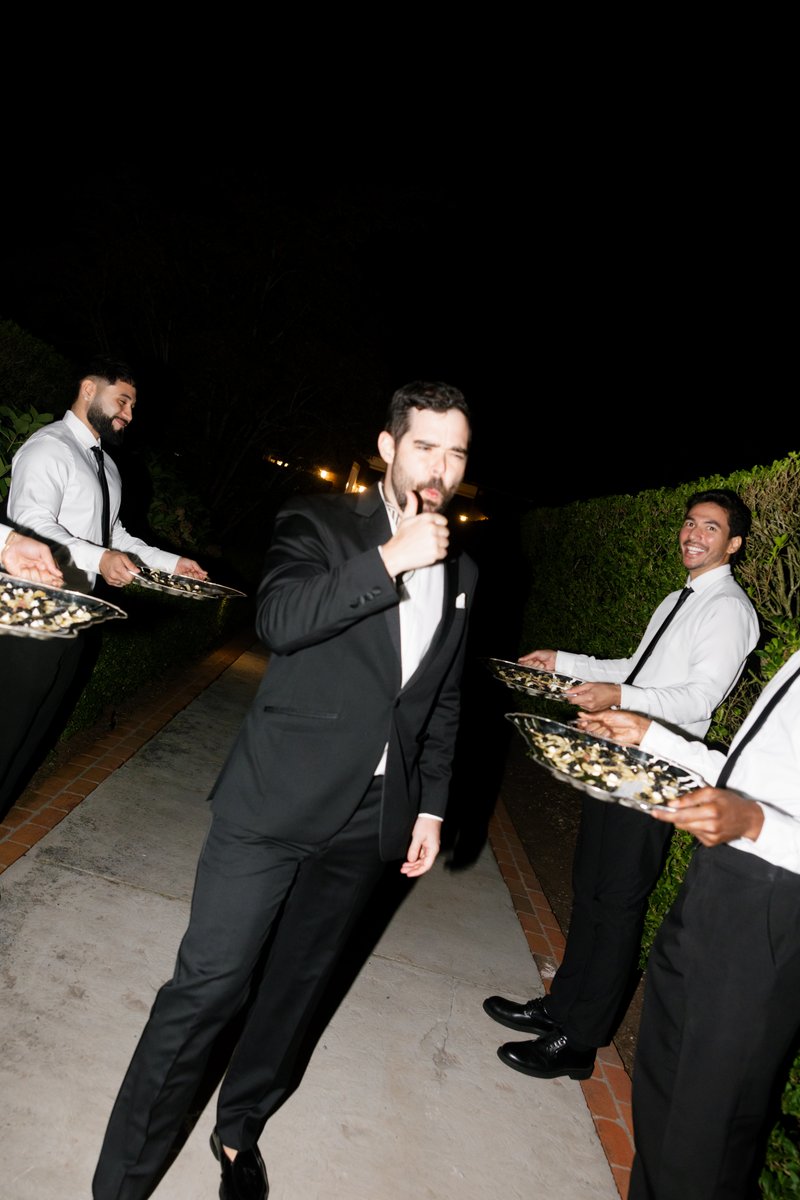 A guest in a tuxedo makes a playful face and gives a thumbs up, interacting with servers carrying trays of appetizers. T
