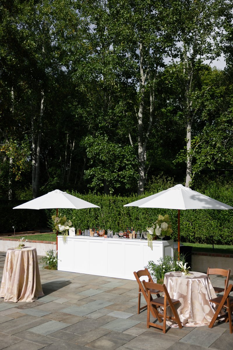An elegant outdoor bar setup is featured on a stone patio, flanked by two white umbrellas and lush green foliage. Nearby