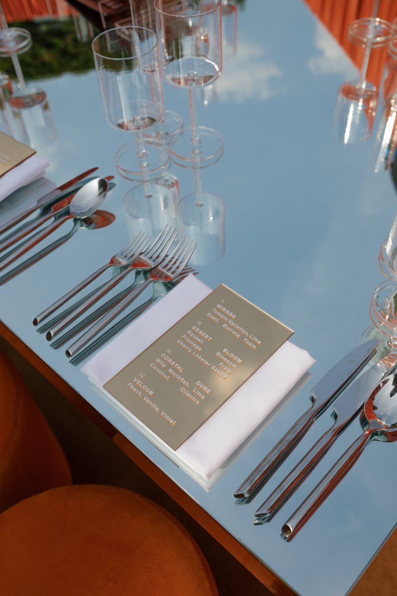 A close-up shot captures a modern and elegant table setting on a highly reflective, light blue surface. Polished silverw