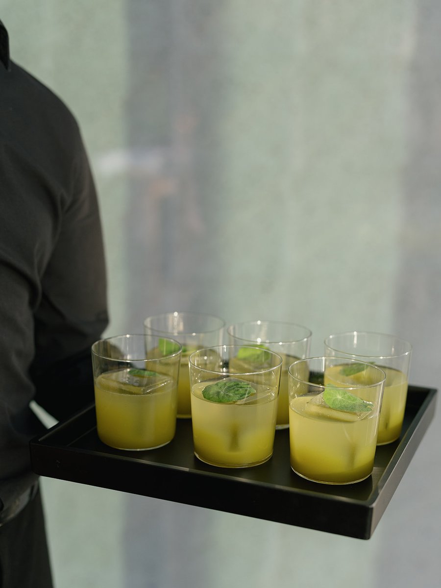 A server, partially visible in a dark uniform, carries a black tray with six refreshing green cocktails. Each drink is g