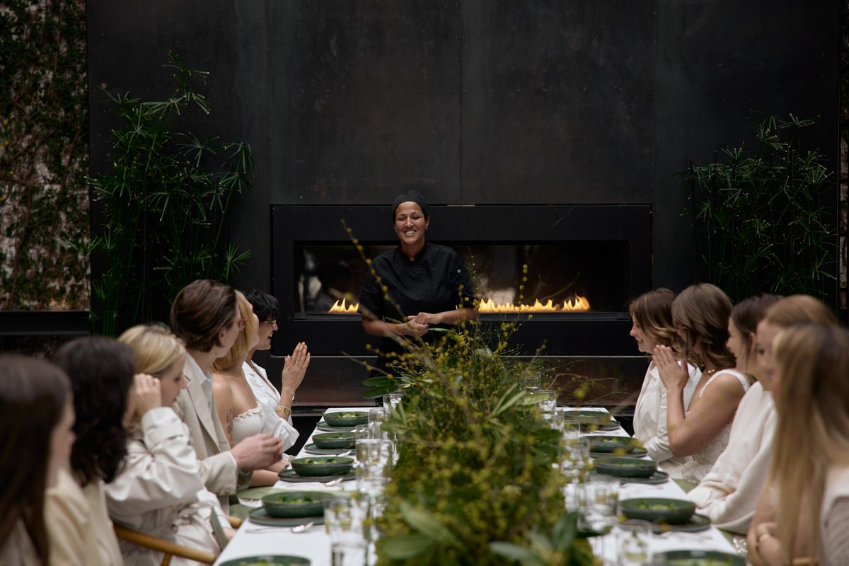 A smiling chef in a black uniform stands in front of a modern fireplace, addressing a group of guests seated at a long, 