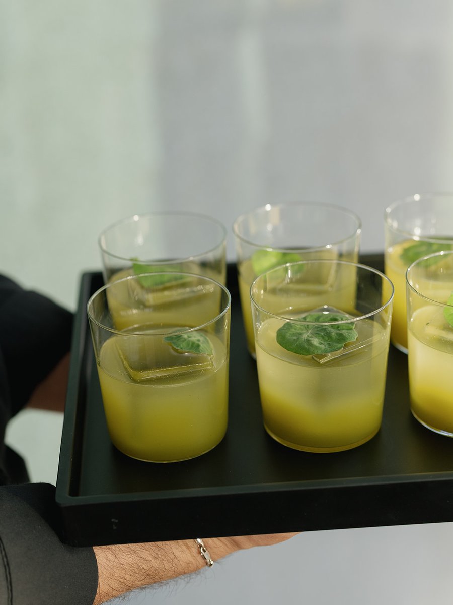 A server carries a black tray laden with several clear glasses, each containing a vibrant green-yellow cocktail. The dri