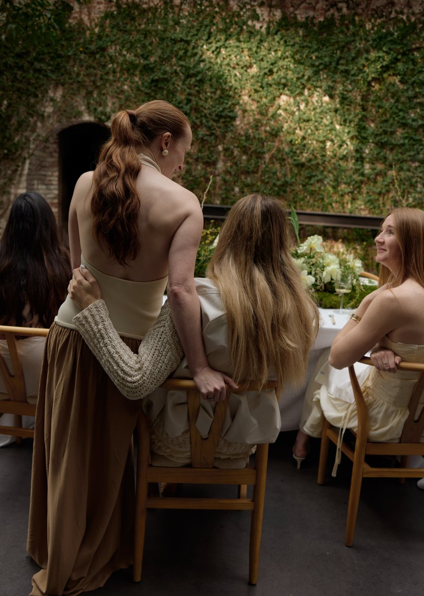 A candid medium-shot captures several women at a table in an intimate setting. One woman with red hair stands, gently em