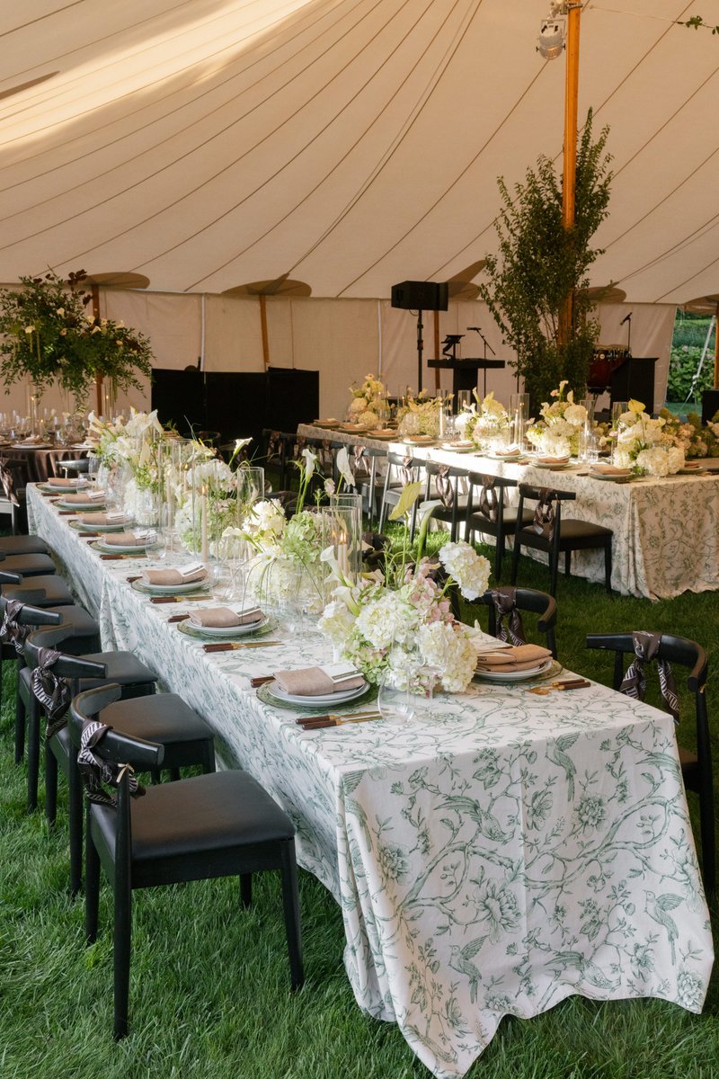 An elegantly set long dining table under a large white tent on a grassy lawn. The table features a distinctive green and
