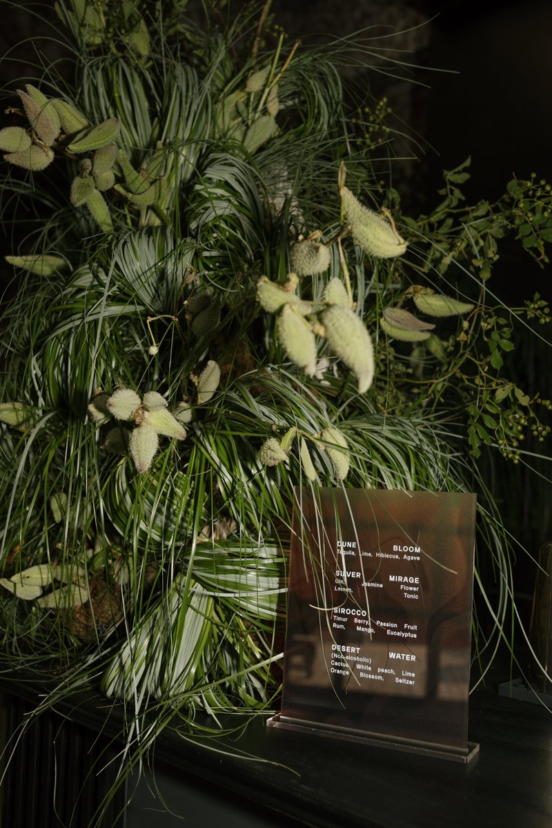 A close-up shot features a stylish, translucent bronze-toned drink menu sign nestled amidst lush, spiky green foliage an