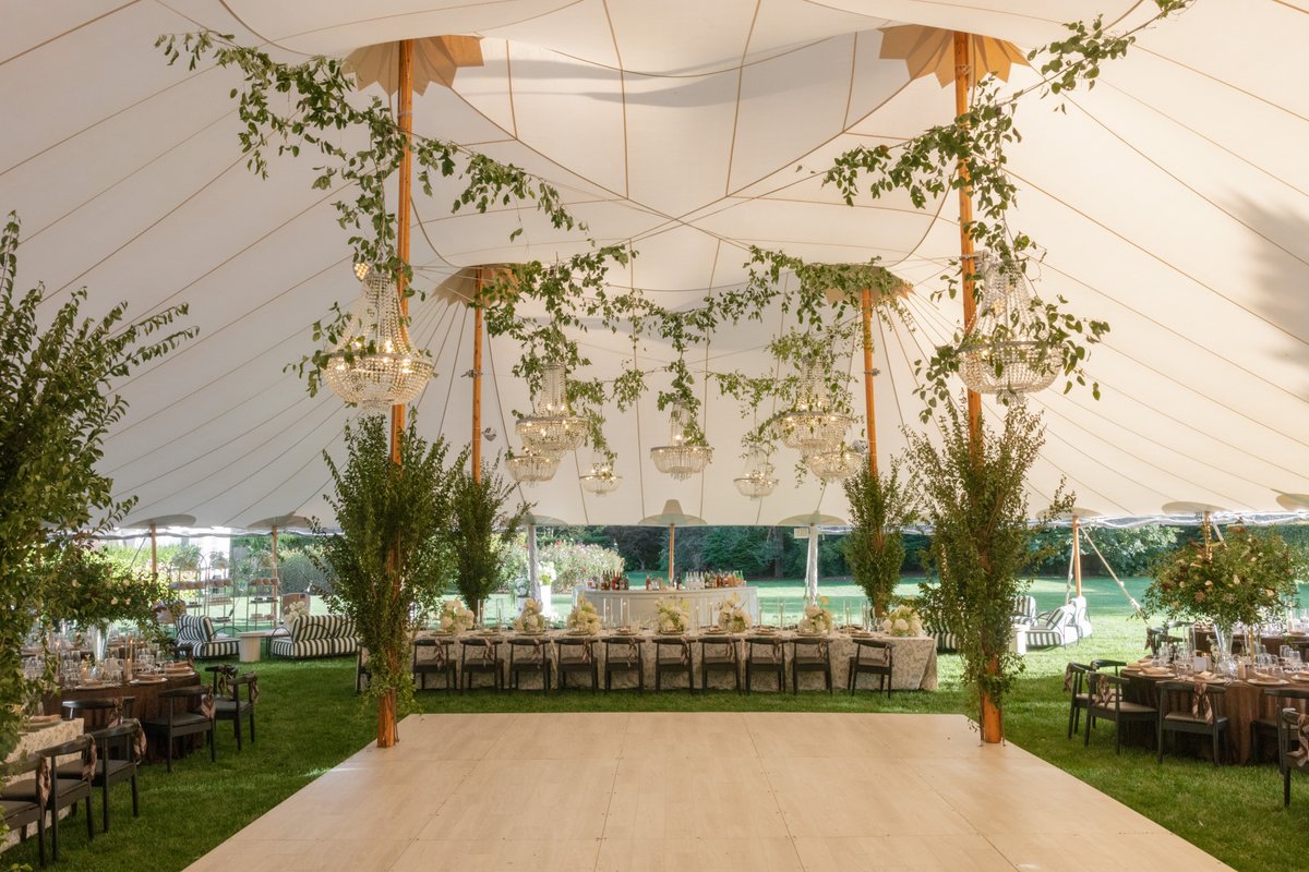 This wide shot captures the interior of a large, elegant white tent set up on a lush green lawn for an event. Ornate cha