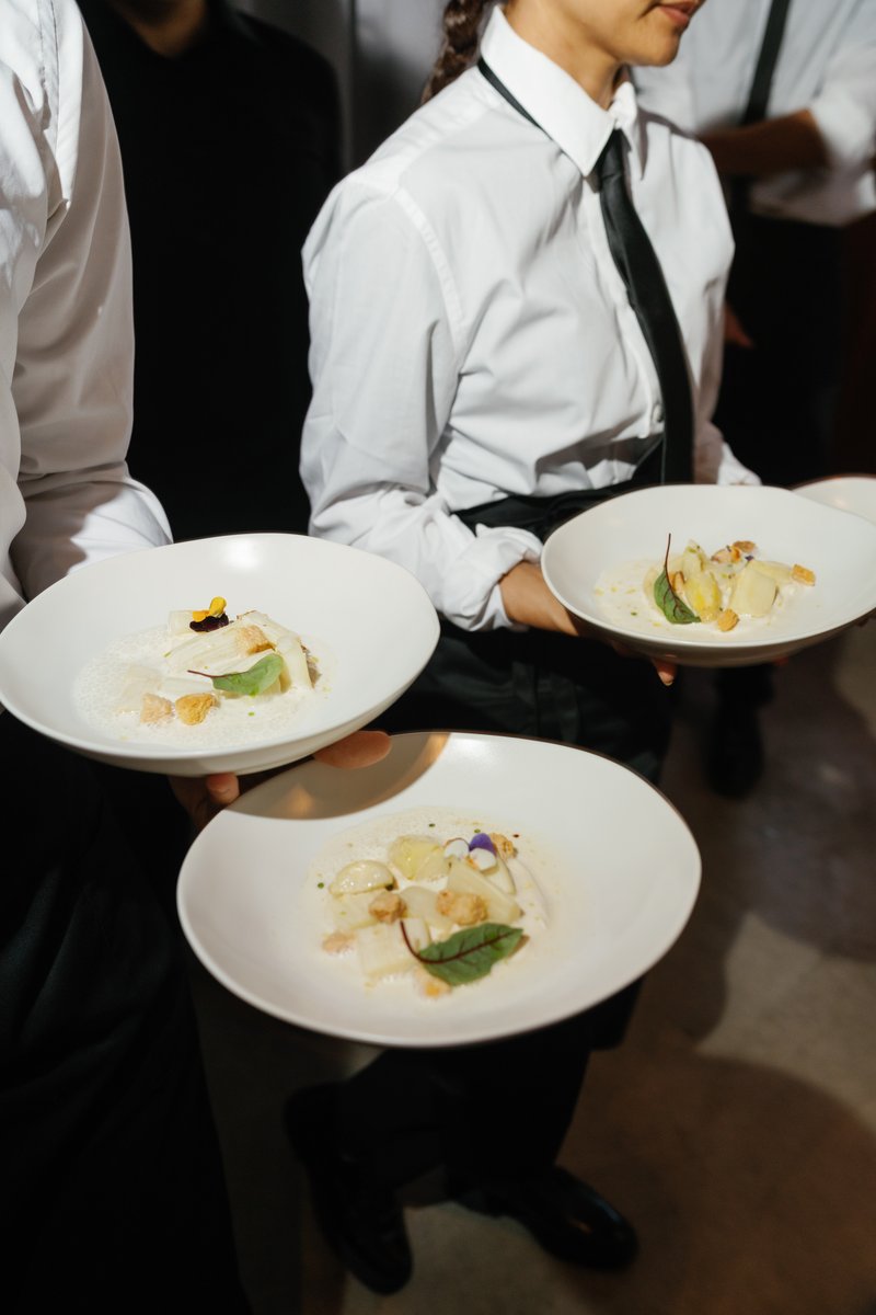 Two servers, dressed in crisp white shirts and black ties/aprons, are captured in motion, carrying multiple elegantly pl