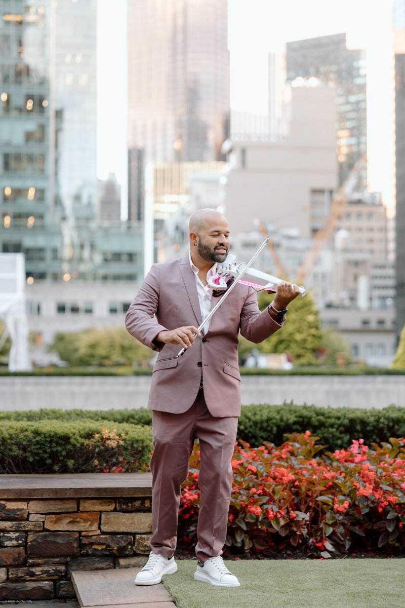 A male musician in a stylish mauve suit plays a white electric violin with pink accents on an outdoor terrace. He is smi