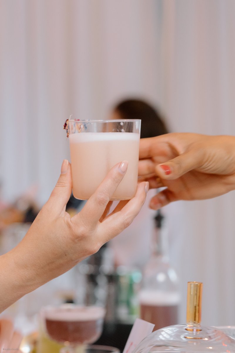 A close-up shot captures two hands exchanging a light pink cocktail, garnished with small dried flowers, at what appears