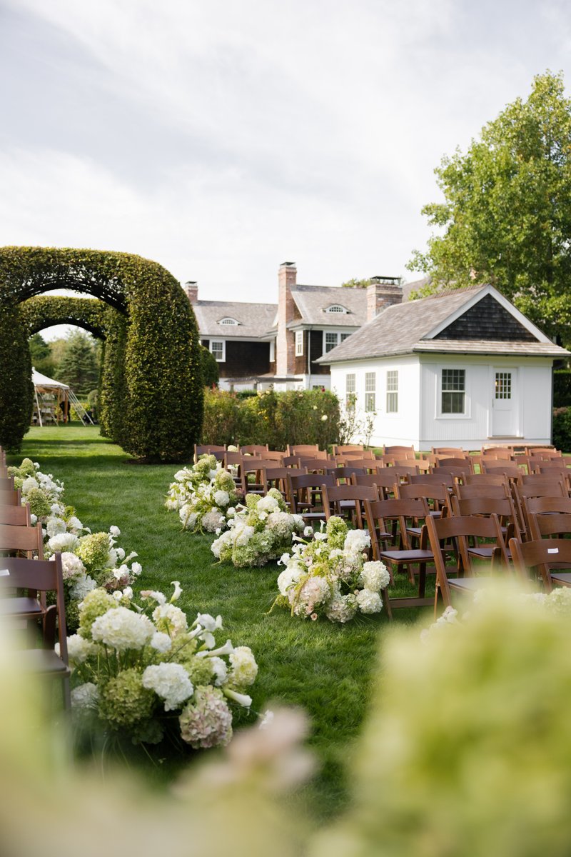 An outdoor wedding ceremony aisle is beautifully set with rows of wooden chairs flanking lush arrangements of white and 
