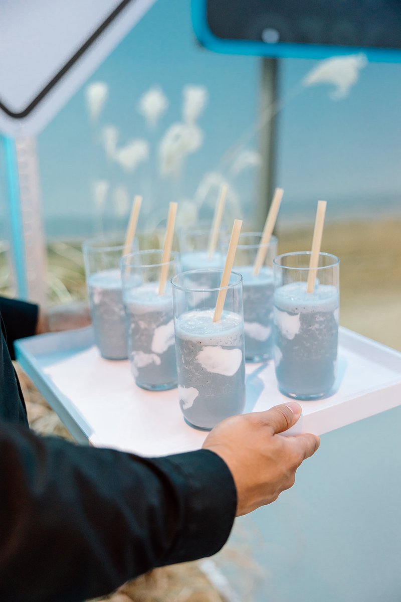 A server carries a white tray filled with several glasses of an intriguing blue-grey beverage, each featuring white, clo