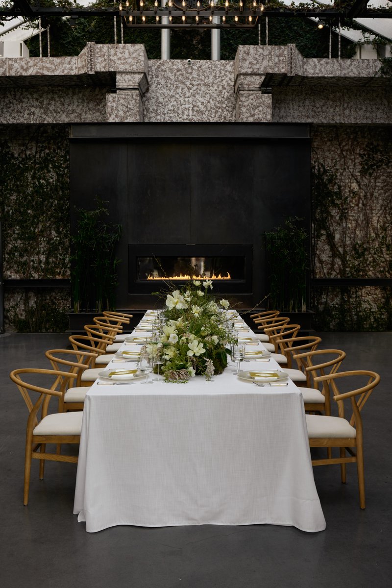 A beautifully set long dining table with a crisp white tablecloth and light wooden chairs, featuring an elaborate green 