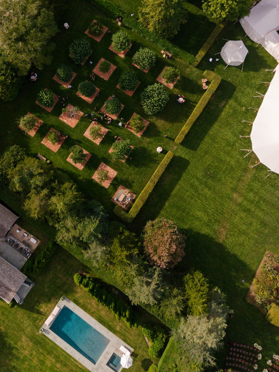 An aerial view captures an elegant outdoor event set within a sprawling, manicured garden. Guests are seen mingling amon