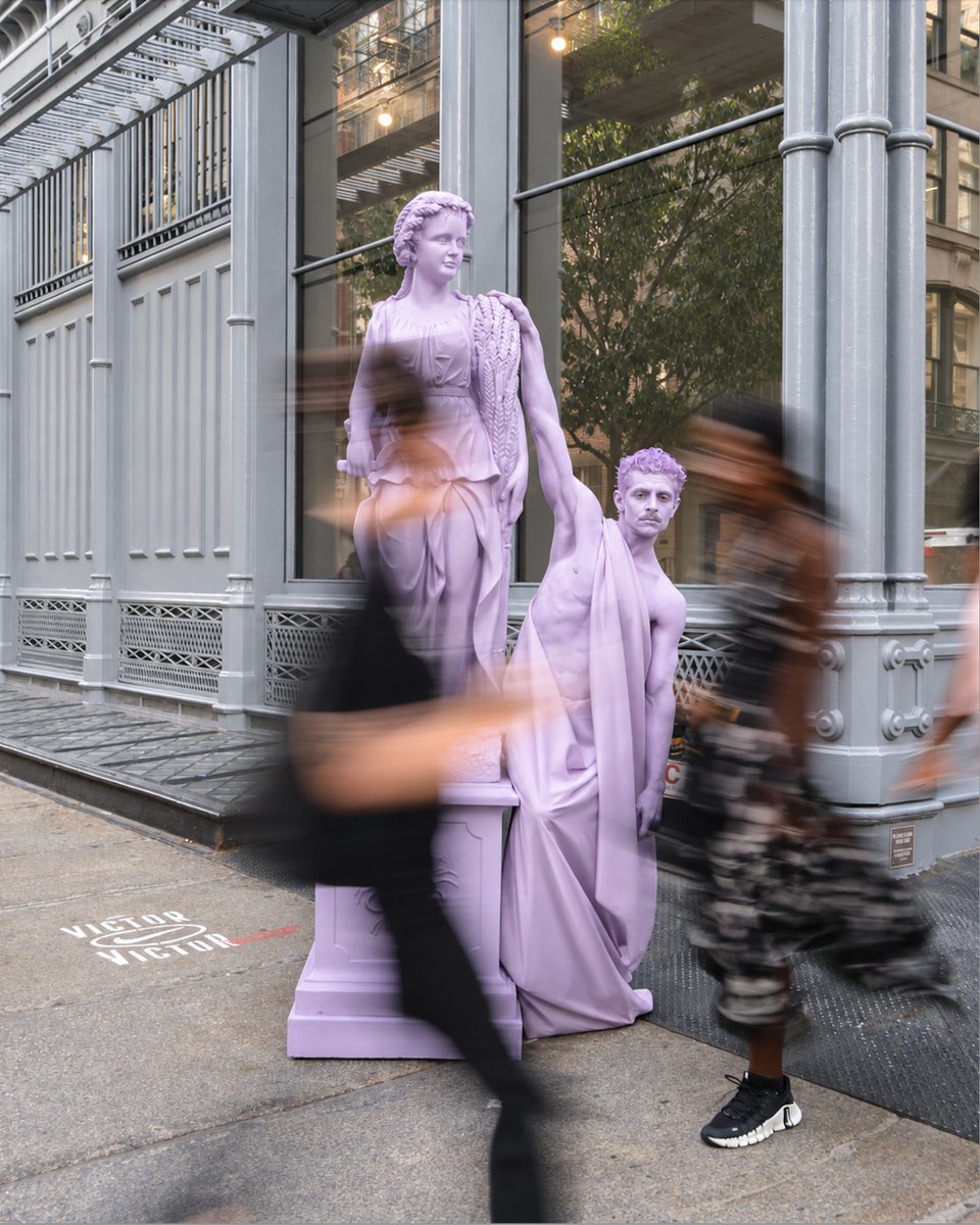 Two lavender-colored statues, one of a woman and one of a man, stand prominently on a city sidewalk. Blurred figures wal