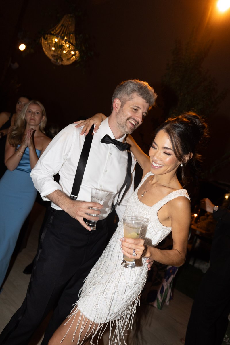 A happy couple is captured mid-dance, laughing and holding drinks, at a lively event. They are surrounded by other guest
