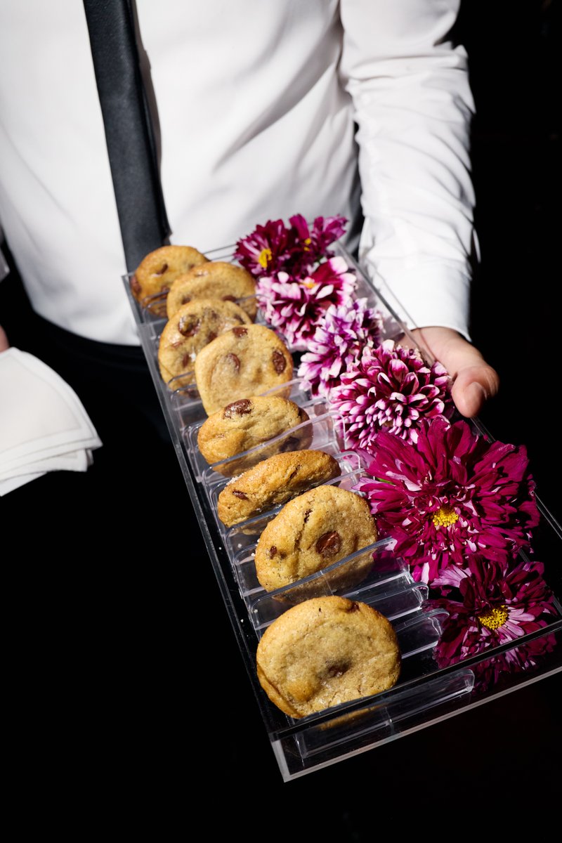 A server, dressed in a white shirt and black tie, holds a clear acrylic tray presenting several chocolate chip cookies a