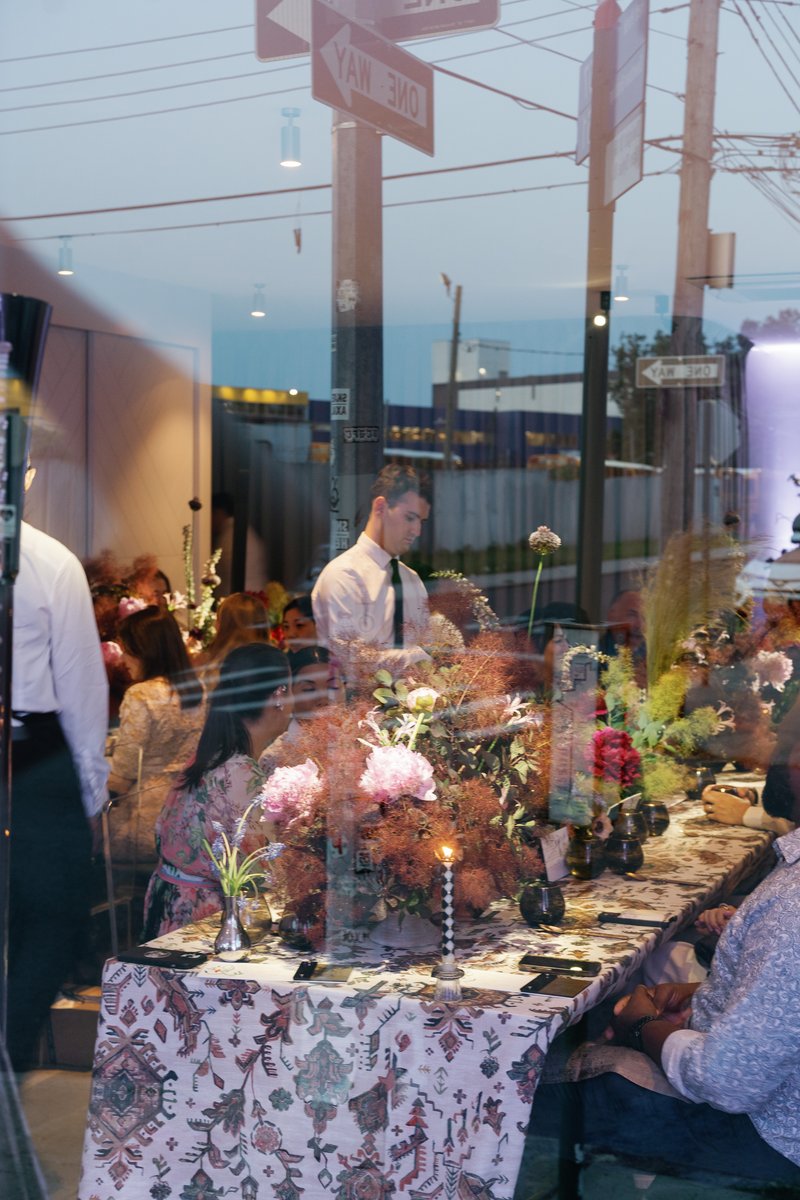 A server in a white shirt and tie attends to a long, elegantly decorated table, seen through a window with street reflec
