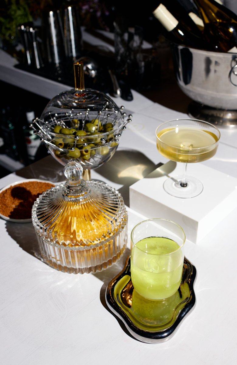 A stylish bar setup features a glass bowl of green olives with picks, a crystal jar of what appears to be dried fruit or