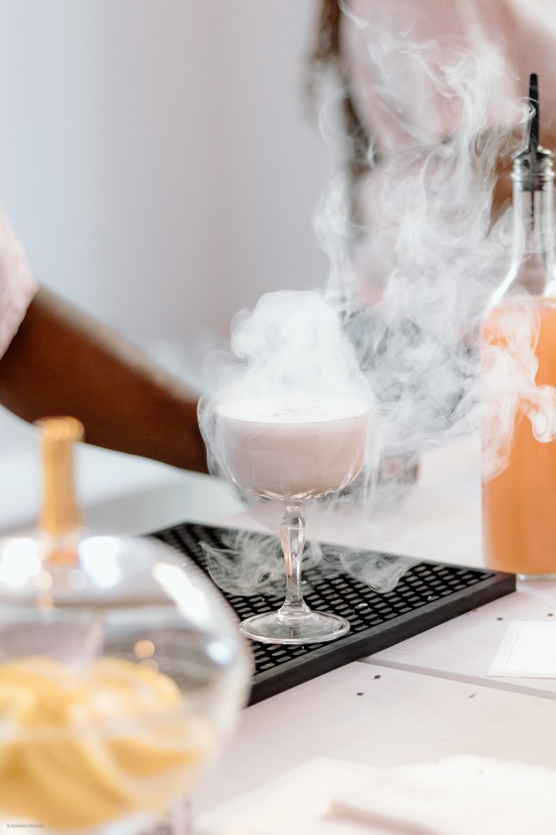 A dramatic close-up captures a smoking pink cocktail in an elegant coupe glass, resting on a black bar mat. The arm of a