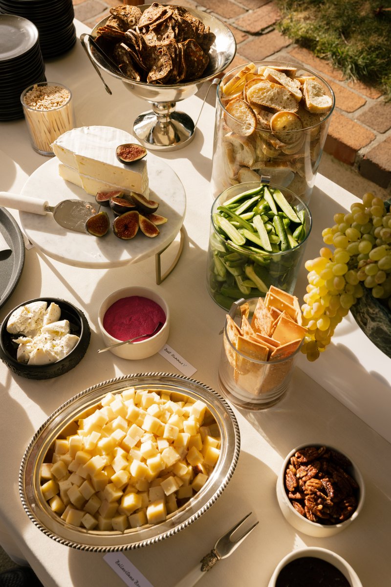 This photo showcases a beautifully arranged cheese and charcuterie spread on a white tablecloth, set outdoors. Various c