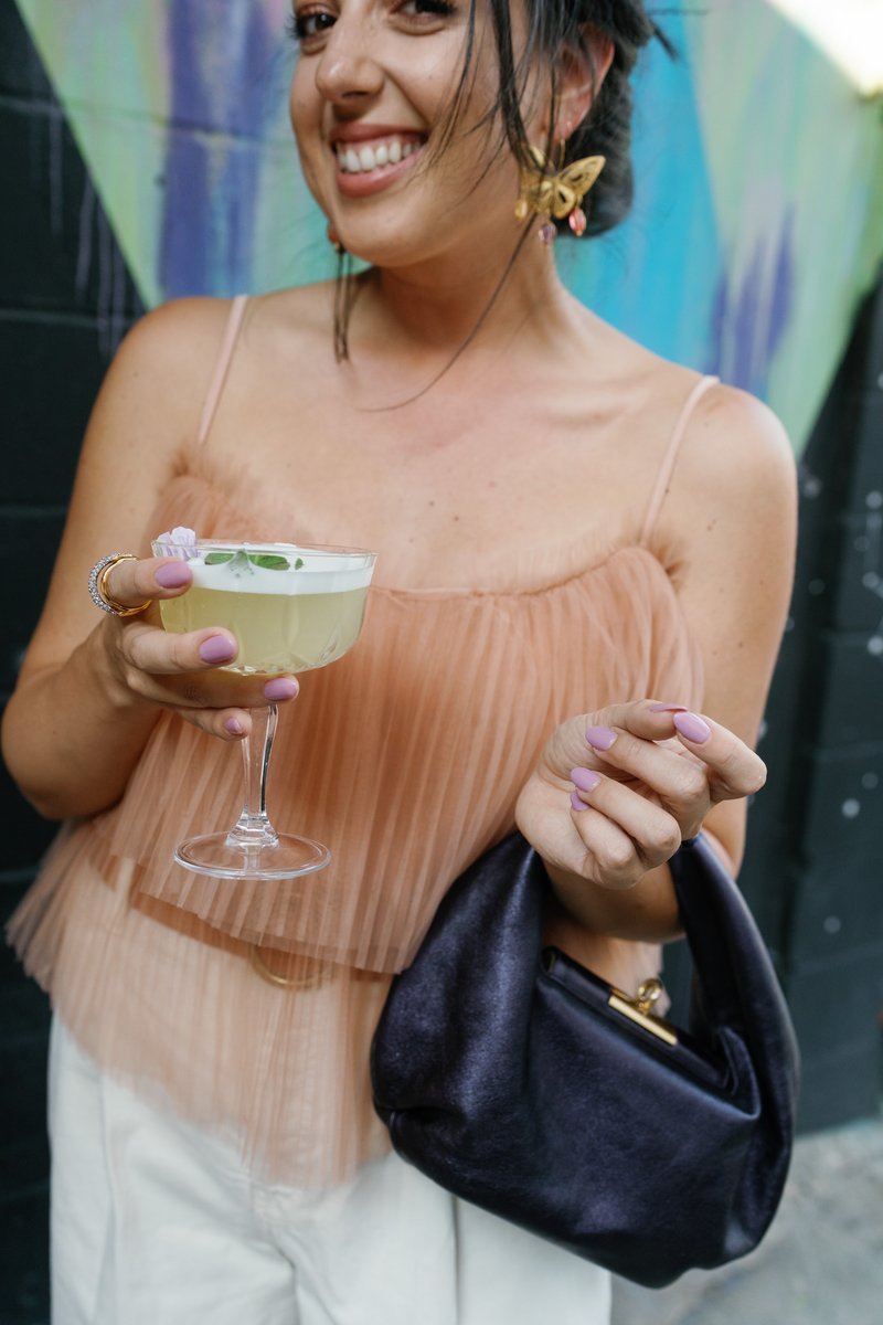 A smiling woman, likely a guest, holds a vibrant cocktail in a coupe glass, garnished with a small flower and mint leave